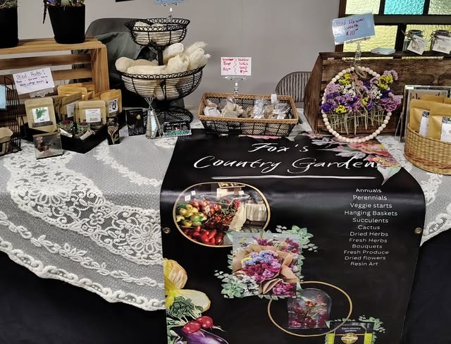 The Fox's Country Garden market table. A black banner printed with the farm name in white script lists the twelve product categories down one side, and the table in front holds packets of dried herbs, a dried flower hoop and bunches of bouquets on a lace cloth.