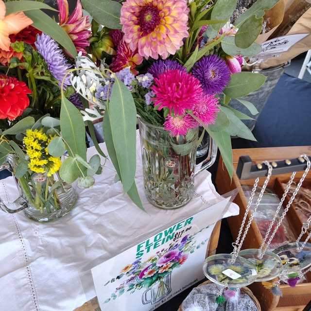 Cut flower arrangements in glass steins on a lace cloth. A large pink and yellow dinnerplate dahlia sits above magenta and purple asters, coral lilies and eucalyptus. On a wooden tray to the side, resin pendants on silver chains with real pressed flowers set inside them.